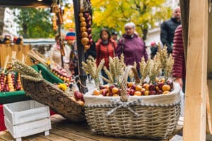 A woven basket filled with peppers, apples, and wheat stalks sits on a wooden stall while autumn crowds browse a lively market in the background.