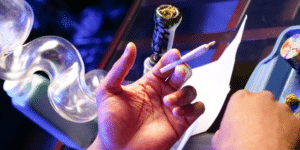 hand holding cannabis pre-roll with glass bong in background